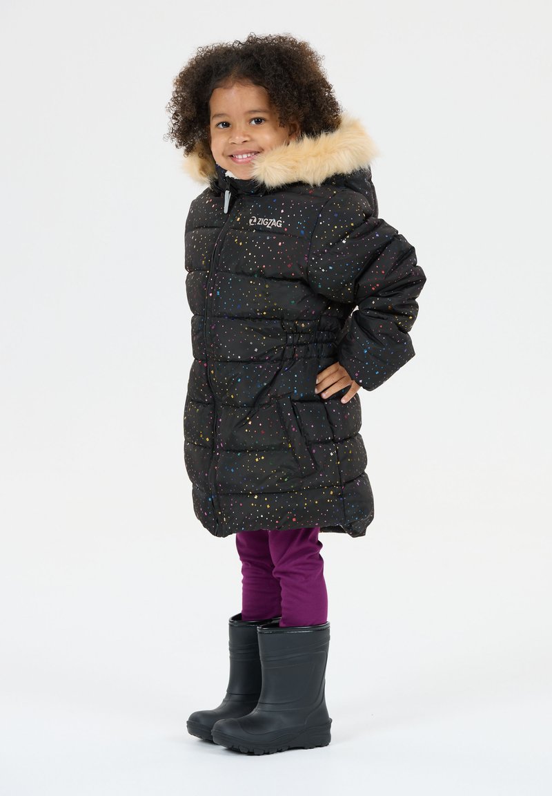 Lille barn smilende, iført en sort puffjakke med multicolor prikker, beige pelshætte, lilla leggings og sorte gummistøvler.