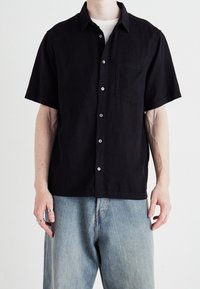Camisa corta de manga negra hecha de tela texturizada, con un bolsillo frontal, cierre de botones y un cuello clásico. Combinada con unos vaqueros de color azul claro.
