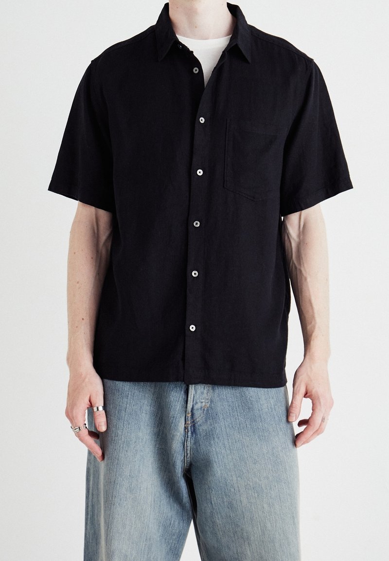 Camisa corta de manga negra hecha de tela texturizada, con un bolsillo frontal, cierre de botones y un cuello clásico. Combinada con unos vaqueros de color azul claro.