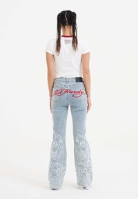 En kvinde står med ryggen til kameraet iført en hvid t-shirt og unikke, dekorative blue jeans med fiske-design og "Ed Hardy" tekst.