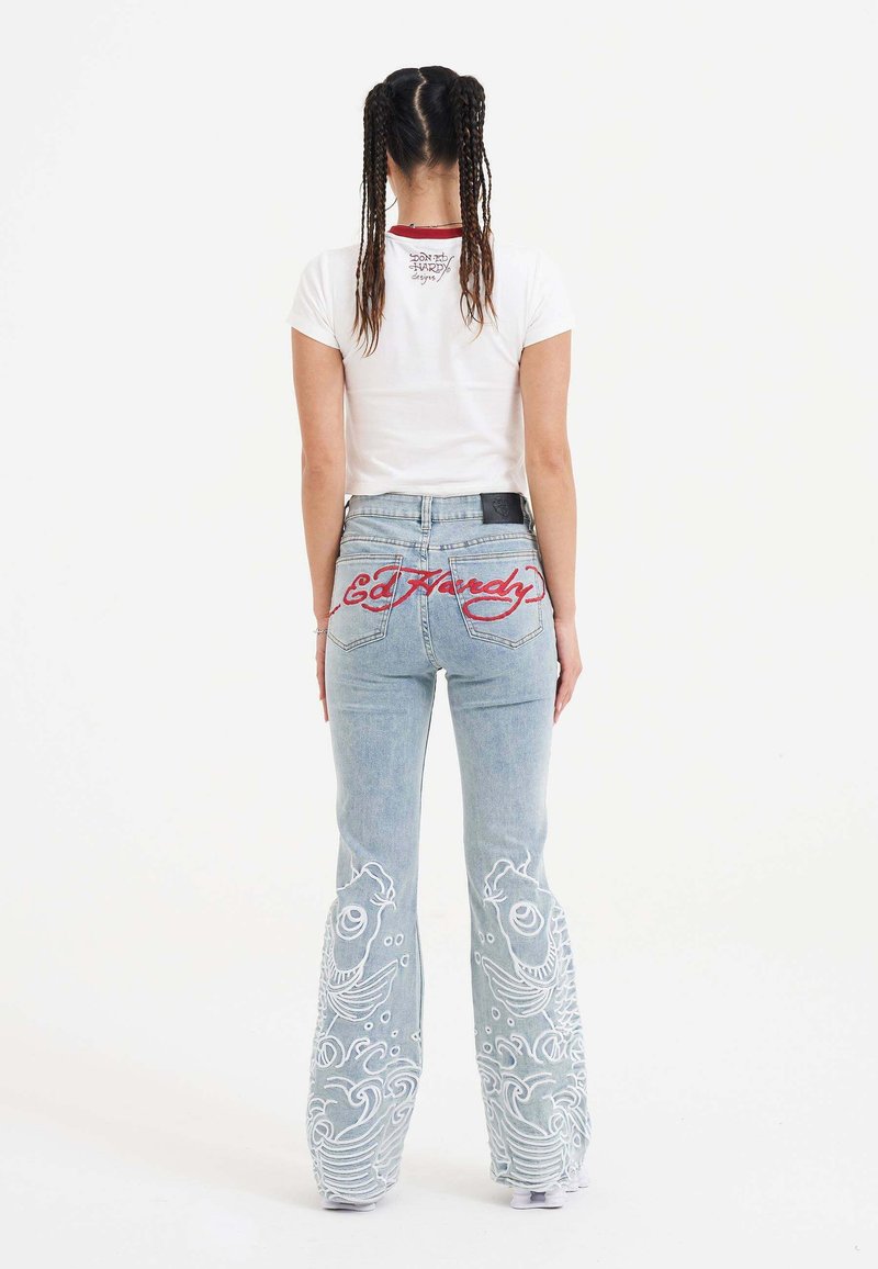En kvinde står med ryggen til kameraet iført en hvid t-shirt og unikke, dekorative blue jeans med fiske-design og "Ed Hardy" tekst.