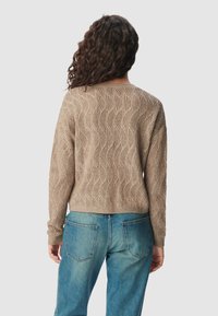 Frau mit lockigem, dunklem Haar, die einen beigen Strickpullover und blaue Jeans trägt und mit dem Rücken zu einem schlichten, hellen Hintergrund steht.