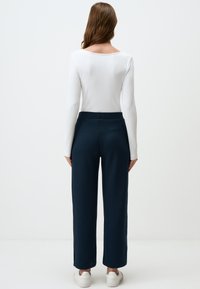 Witte lange mouwen top met een geribbelde textuur gecombineerd met donkerblauwe wijde broek met een subtiel patroon. Model draagt witte sneakers.