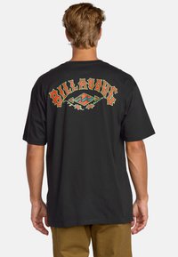 Camiseta negra con el texto colorido "Billabong" acompañado de acentos florales y un gráfico de olas en la parte trasera. Manga corta, ajuste estándar.