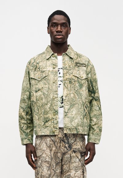 Πράσινο καμουφλάζ μπουφάν με κούμπωμα με κουμπιά, δύο μπροστινές τσέπες και λογότυπο. Φορεμένο πάνω από λευκό μπλουζάκι με γραφικά και παντελόνι με σχέδια. Υφασμένο ύφασμα.