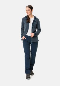 Un hoodie zippé marine avec des accents bleu clair, associé à un pantalon marine. Le modèle porte une chemise blanche en dessous. Des chaussures d'extérieur complètent le look.