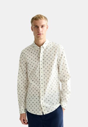 Uomo biondo che indossa una camicia color avorio con piccoli motivi di sole e cerchi, abbinata a pantaloni blu scuro, in piedi davanti a uno sfondo semplice.