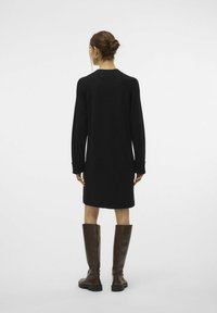 Schwarzes Strickkleid mit langen Ärmeln, midi-langer Schnitt, strukturierten Stoff und umgeschlagenen Bündchen; kombiniert mit braunen kniehohen Stiefeln.