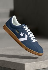 Zapatilla baja en azul marino con parte superior de gamuza, logo de estrella blanca, cordones blancos y suela de goma. Presenta detalles de costura mínimos.