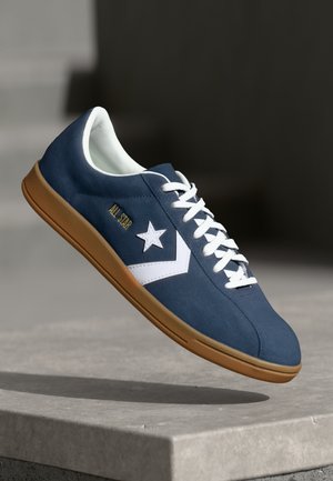 Navy blue low-top sneaker with a suede upper, white star logo, white laces, and a gum rubber sole. Features minimal stitching details.