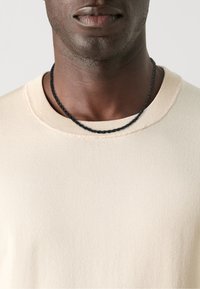 Beige bordás pulóver kerek nyakkal, fekete fonott nyaklánccal párosítva. Közeli felvétel, amely a szövet textúrájára és részleteire összpontosít.