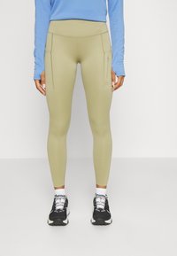 Világos olívazöld leggings, magas derékkal, két oldalsó zsebbel és sima, rugalmas anyagból készült. Fekete sportcipővel párosítva.