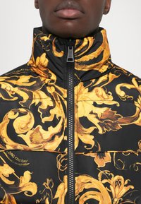 Veste matelassée noire ornée de motifs floraux dorés complexes, col haut et fermeture éclair centrale avec des détails du logo sur le tissu.