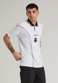 Hvid polo-skjorte med sort krave og røde accenter. Udstyret med et logo på brystet og detaljering af syninger. Lavet af glat stof.