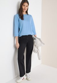 Femme portant un haut à manches longues bleu clair, pantalon noir, baskets blanches, tenant une veste gris clair, debout contre un mur blanc et beige.
