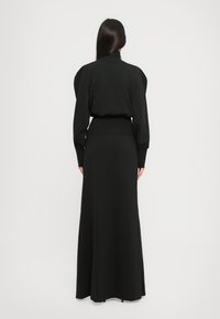 Frau mit langen, dunklen Haaren, die ein langes schwarzes Kleid mit aufgepufften Schultern trägt und vor einem schlichten weißen Hintergrund steht.