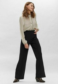 Blusa crema con pois neri e chiusura con bottoni; pantaloni neri a vita alta con gamba larga; modella in una posa casual.