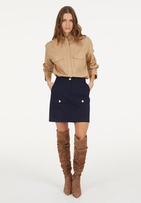 Beige button-up shirt with two pockets, navy mini skirt with gold hardware, and brown knee-high suede boots. Smooth texture, fitted design.
