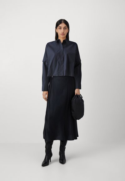 Cropped shirt nera in tessuto morbido, abbinato a una lunga gonna nera in satin e stivali col tacco neri fino al ginocchio. Tiene in mano una borsa nera.