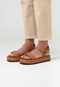 Brown platform sandals with a textured upper, featuring wide straps and adjustable ankle straps. Paired with light beige cropped pants.