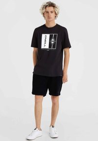 Camiseta de algodón negra con un diseño gráfico en blanco y gris claro, que incluye texto y formas geométricas. Combinada con pantalones cortos de pana oscuros y zapatillas blancas.