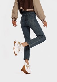 Sneakers bianchi e dorati con una suola testurizzata, abbinati a jeans blu scuro e a una giacca marrone e beige, visti di lato.