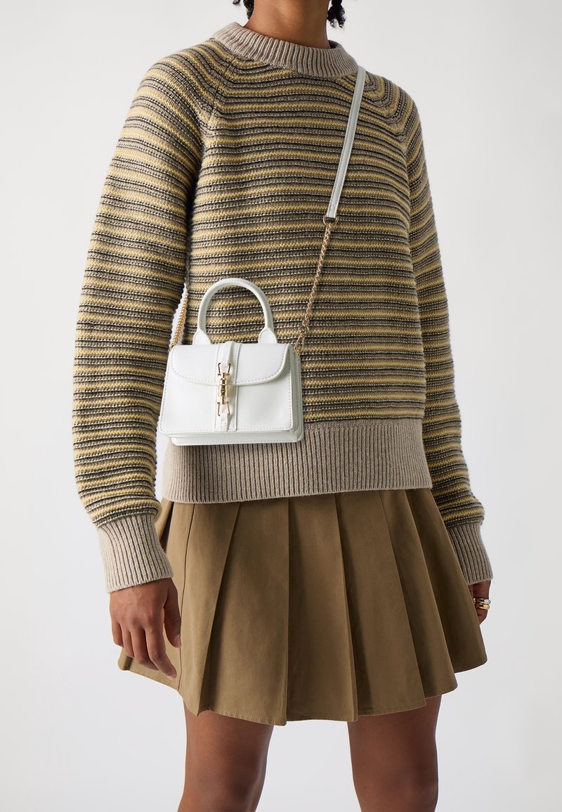 Femme portant un pull rayé à manches longues et une jupe plissée marron avec un petit sac blanc à fermoir doré porté en bandoulière.