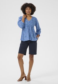 Világoskék, textúrált blúz V kivágással és ráncolt elejével, sötét rövidnadrággal és barna tömbsarkú szandállal párosítva.