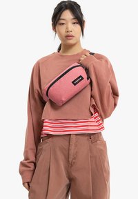 Rosa, glittrig axelremsväska med en svart dragkedja och logotyp, rektangulär form, bärs över en baggy brun sweatshirt med randig nederkant.