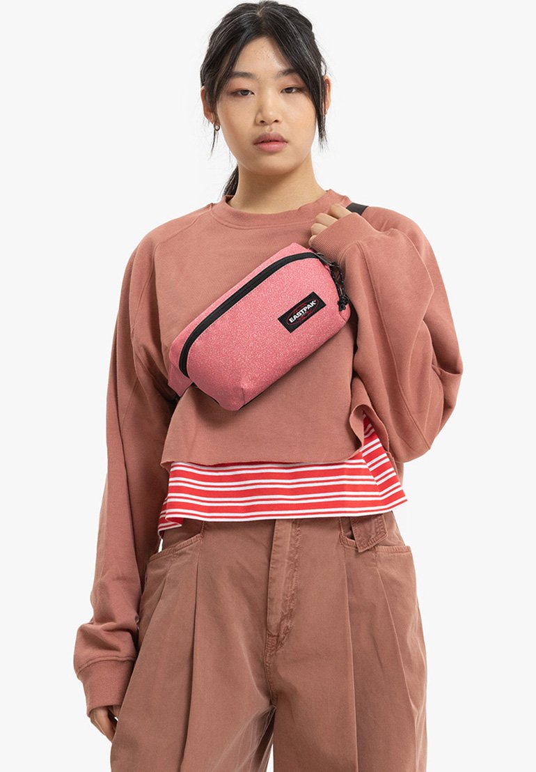 Rosa, glitzernde Umhängetasche mit schwarzem Reißverschluss und Logo, rechteckige Form, getragen über einem lässigen braunen Sweatshirt mit gestreiftem Saum.