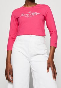 T-shirt rosa a maniche lunghe con scollatura rotonda e stampa logo bianca, abbinato a pantaloni bianchi a vita alta; tessuto di cotone liscio.