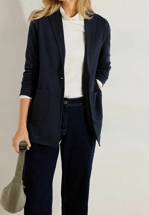 Femme portant un blazer bleu marine et un jean foncé, tenant un petit sac à main beige, debout devant un fond clair uni.
