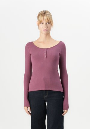 Kvinne med blondt hår iført en mauve ribbet langermet topp med knapper og mørkeblå jeans, som står foran en hvit bakgrunn.