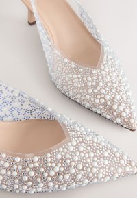 Escarpins transparents à bout pointu décorés de perles blanches de tailles variées et de petits strass clairs sur un fond neutre.