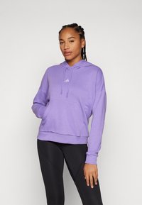 Fialová mikina s kapucňou, prednou vreckom a logom Adidas; má mäkkú látku a voľný strih; skombinovaná s čiernymi legínami.