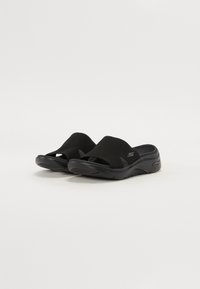 Svarte slidesandaler med mesh-overdel, åpen tådesign, teksturert fotseng og polstret såle. Merkelig logo på siden.