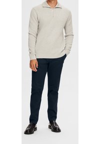 Hellgrauer Strickpullover mit einem halben Reißverschluss, kombiniert mit marineblauer Hose und schwarzen Loafers, für ein sauberes, neutrales Outfit.