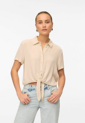 Blouse beige à manches courtes avec un col et un nœud à l'avant, fabriquée en tissu léger. Associée à un jean taille haute bleu clair.
