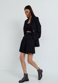 Zwarte op maat gemaakte blazer met scherpe schouders, gecombineerd met een cropped top en hoge, geplooide shorts. Glanzende donkere enkellaarsjes maken de look compleet.