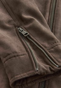 Chaqueta de tela marrón y suave con cremallera frontal y bolsillos con cremallera lateral. Presenta herrajes en tono plateado y una textura lisa.