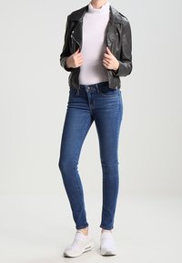 Chaqueta de cuero negro estilo biker, suéter de cuello alto blanco, jeans skinny azules y zapatillas deportivas blancas. Modelo de pie con las manos sobre la chaqueta, fondo neutro.