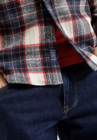 Czerwono-czarna koszula flanelowa w kratę, z bawełnianego materiału, z wzorem w jodełkę. Nosi się na czerwonej koszulce, w zestawie z ciemnoniebieskimi dżinsami.