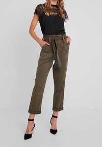 Top in pizzo nero con scollatura rotonda, abbinato a pantaloni cargo verdi oliva con cintura, tasche laterali e orli risvoltati. Scarpe nere con tacco alto.