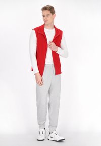 Rote Fleece-Weste mit einem Reißverschluss vorne über einem weißen Langarm-Shirt. Kombiniert mit hellgrauen Sweatpants und Sneakers mit schwarzen Akzenten.
