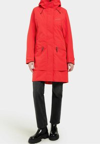 Röd vattentät parkas med luva, dragkedja framtill, två framfickor och justerbara dragsnören. Kombinerad med svarta raka byxor och stövlar.