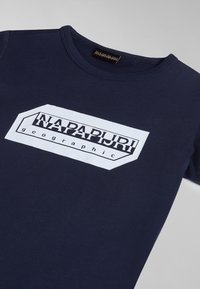 Tummansininen puuvillapaita, jossa on valkoinen suorakulmainen logomuotoilu, jossa lukee "NAPAPIJRI" ja "geographic" mustalla. Klassinen pyöreä kaula-aukko.