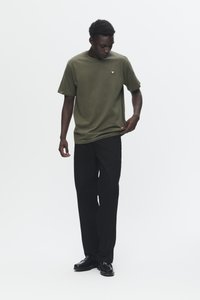 Man wearing olive green t-shirt with small emblem, black pants, and black shoes, standing and looking down while holding shirt hem.