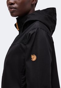 Person wearing a black hooded jacket with a tan fox logo patch on the upper sleeve, shown from the side against a plain background.
