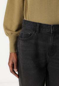 Jean en denim noir avec une texture mate, design à cinq poches et fermeture à bouton standard. Associé à un pull ample vert olive.