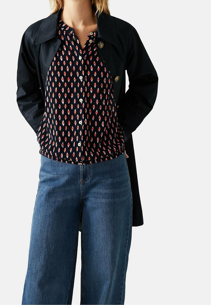 Impermeabile navy sopra una blusa a motivi in blu scuro con forme romboidali rosse. I jeans sono larghi, in denim di lavaggio medio.